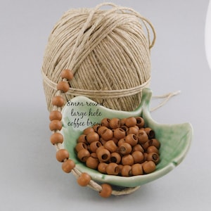 May include: A close-up of a bowl of 8mm round wooden beads with a large hole. The beads are a coffee brown color. The beads are in a small green bowl. There is a ball of twine in the background.