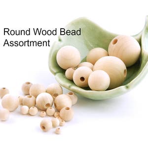 May include: A collection of round, unfinished wood beads in various sizes, some with pre-drilled holes. The beads are displayed in a light green, leaf-shaped dish and scattered on a white surface. Text on the image reads "Round Wood Bead Assortment."