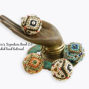 Op de afbeelding: Vier kralenbollen met ingewikkelde geometrische patronen in tinten zwart, wit, goud, rood, groen, blauw en turquoise. De kralen worden getoond op een bronzen handbeeld.