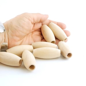 May include: A collection of unfinished wooden oval beads with a hole drilled through the center. The beads are a light beige color and are smooth. The beads are scattered on a white surface.
