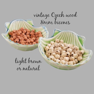 May include: Two small bowls filled with vintage Czech wood 8mm bicones. One bowl contains light brown beads and the other contains natural wood beads.