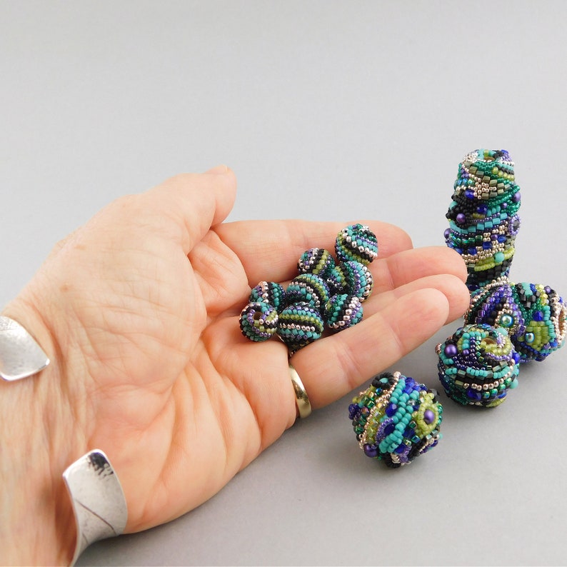 May include: A hand holding a collection of small, colorful, beaded spheres. The beads are a mix of green, blue, purple, and black. The beads are arranged in a pile on the palm of the hand.