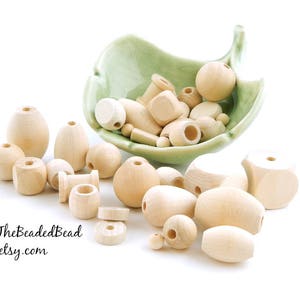 May include: A collection of natural wood beads in various shapes and sizes, including round, oval, and square beads. The beads are displayed in a green ceramic bowl and on a white surface. The text "TheBeadedBead.etsy.com" is visible in the bottom left corner of the image.