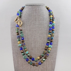 Puede incluir: Un collar de dos hilos con cuentas azules, marrones y verdes. El collar tiene un cierre dorado con un diseño de hoja.
