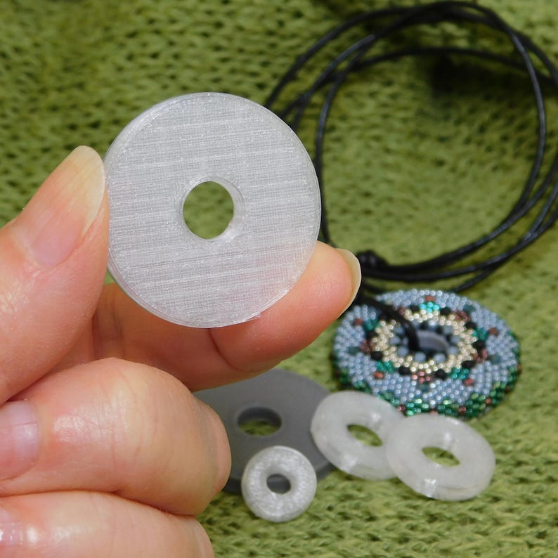 Hand holding a large 1.25 inch black translucent crystal washer alongside smaller 3/4 inch and 1/2 inch 3D printed silver and crystal discs to demonstrate relative scale.
