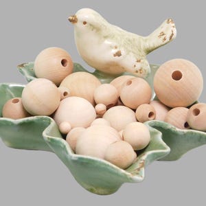 Puede incluir: Un plato de cerámica verde claro con forma de hoja y una figura de pájaro en el borde. El plato está lleno de una colección de cuentas de madera redondas y sin tratar de varios tamaños, cada una con un orificio central para ensartar.