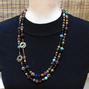 Puede incluir: Un collar de múltiples hilos con cuentas marrones, doradas, turquesas y azules. El collar presenta dos dijes de metal con un diseño geométrico.