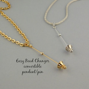 Könnte beinhalten: Zwei Halsketten mit Anhänger-/Pin-Aufsätzen. Eine ist goldfarben mit einer Kette, die andere silberfarben mit einem Band. Der goldene Anhänger trägt den Text "Easy Bead Changer convertible pendant/pin". Die Anhänger haben einen langen Stift mit Perlen und ein dekoratives Ende.