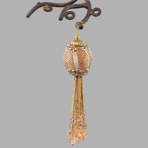 Puede incluir: Un adorno de borla de cuentas con una esfera de color dorado y melocotón y una larga borla de cuentas doradas y melocotón.