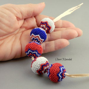 May include: A strand of beaded necklace with red, white, and blue beads. Each bead is a sphere with a geometric pattern. The beads are strung on a thin white cord. The text "Sharri Moroshok" is visible in the image.
