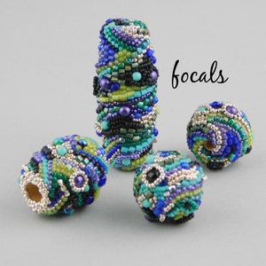 May include: Four colorful beaded focal beads in shades of blue, green, black, and silver. The beads are round and oval shaped and have a variety of textures. The word "focals" is written in black ink on a white background.
