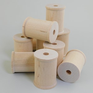 May include: A collection of light-colored, unfinished wooden spools stacked together. Each spool features a cylindrical body with a central hole and raised edges at the top and bottom, showcasing the natural grain and texture of the wood material.