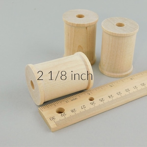 May include: Three unfinished wooden spools, each measuring 2 1/8 inches in diameter, are arranged on a wooden ruler. The ruler is marked in inches and 1/8 inch increments.