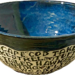 May include: A handmade ceramic bowl with a vibrant blue interior and a dark green and black rim. The exterior features a cream-colored band with black text that reads "CAPE COD TAKES KESTREL TUFTED HAWK FLYING WOODS".