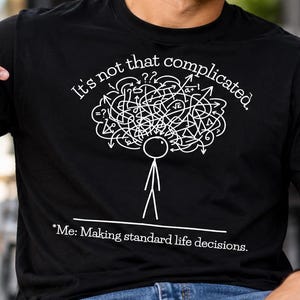 May include: A black short-sleeved t-shirt featuring a white graphic of a stick figure with a tangled mess of lines and mathematical symbols above its head. The text reads, "It's not that complicated." and "*Me: Making standard life decisions."