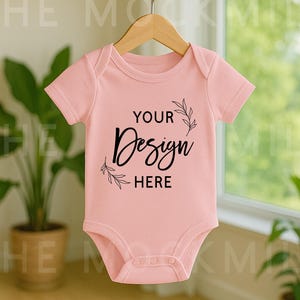 Op de afbeelding: Een lichtroze babybody met korte mouwen hangt aan een houten hanger. De body heeft de tekst "YOUR Design HERE" in zwart schrift. Het ontwerp wordt geflankeerd door bladillustraties.