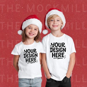 May include: Two children wearing white t-shirts with the text "YOUR DESIGN HERE" in black, and Santa hats with red and white trim. The background is a solid red color.