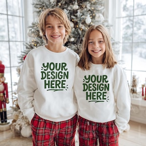 May include: Two children wearing white sweatshirts with the text "YOUR DESIGN HERE" in green, paired with red plaid pajama pants. They are smiling in front of a decorated Christmas tree, creating a festive holiday scene.