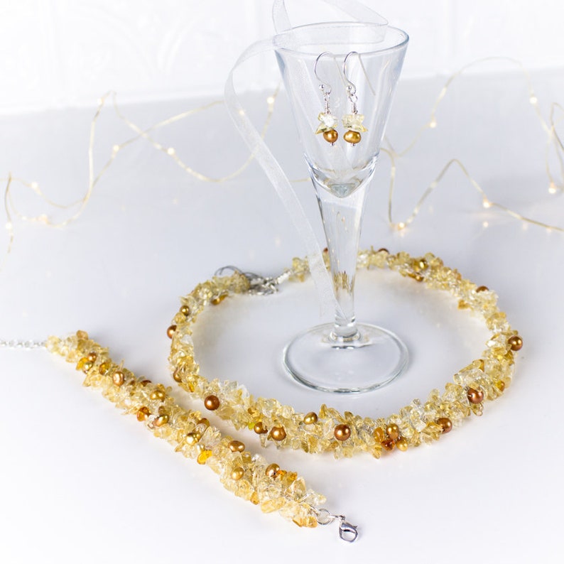K&ouml;nnte beinhalten: Ein Schmuckset aus Citrin-Edelsteinen mit einer Halskette, einem Armband und Ohrringen. Die Halskette und das Armband bestehen aus kleinen, unregelm&auml;&szlig;igen Citrin-St&uuml;cken und kleinen goldenen Perlen. Die Ohrringe sind silberfarben mit kleinen Citrin-Steinen.