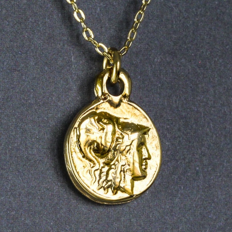 Peut inclure: Un collier en or avec un pendentif rond repr&eacute;sentant un profil d&eacute;taill&eacute; d'une t&ecirc;te de femme portant un casque. Le pendentif rappelle une ancienne pi&egrave;ce de monnaie grecque.