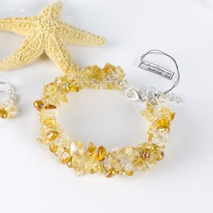 K&ouml;nnte beinhalten: Ein goldfarbenes und klares Kristall-Armband mit einem silbernen Verschluss. Das Armband besteht aus kleinen, unregelm&auml;&szlig;igen Kristallst&uuml;cken und ist mit kleinen goldenen Perlen verziert. Das Armband ist auf einem wei&szlig;en Hintergrund mit einem Seestern abgebildet.