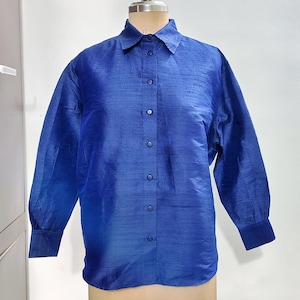Può includere: Una camicia blu brillante con bottoni, maniche lunghe e colletto classico. La camicia è esposta su un manichino, mostrando il suo tessuto liscio e strutturato. I polsini sono di una tonalità di blu più scura.