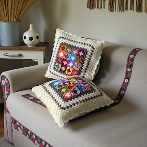 May include: Two square crochet cushions with a colourful patchwork design rest on a beige sofa. The cushions have a cream-coloured base with a scalloped edge and a brown border. The sofa has a floral patterned trim.