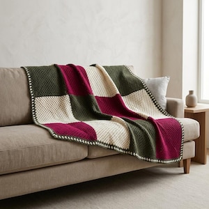 May include: A crochet blanket featuring a checkered pattern of dark olive green, deep magenta, and cream-colored squares. The blanket is constructed with a textured stitch and finished with a cream-colored border along the outer edges.