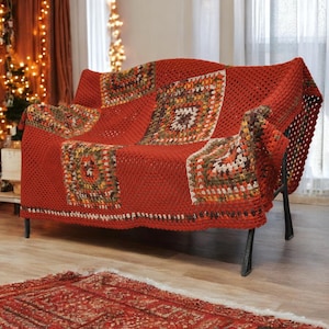 Peut inclure: Une couverture rouge au crochet avec des motifs carrés dans les tons de marron, de vert et de crème recouvre un canapé à cadre en métal noir. La couverture est drapée sur le canapé. Un tapis rouge est posé sur le sol.