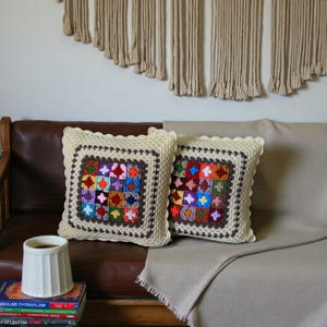May include: Two square crochet cushions with a colourful patchwork design. The cushions have a cream-coloured border and a dark brown inner border. They rest on a brown leather sofa with a beige blanket. A white mug and books are in the foreground.