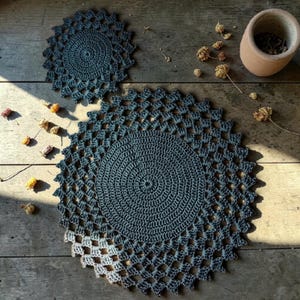 May include: Two circular dark gray crochet doilies featuring a solid center and an openwork lattice border. The larger doily is positioned below the smaller one, both displaying a consistent yarn texture and intricate stitch pattern throughout.