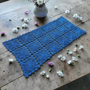 Könnte beinhalten: Handgefertigter, blauer Häkeltischläufer mit geometrischem Muster. Der Läufer ist rechteckig und weist ein sich wiederholendes Rautenmuster auf. Der Läufer liegt auf einem rustikalen Holztisch, der mit kleinen weißen und rosa Blumen und einer kleinen Vase dekoriert ist.