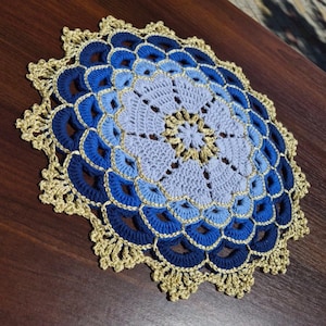 May include: A round, crocheted doily featuring a circular pattern with concentric rings of light blue, medium blue, and dark blue yarn. The outer edge is finished with a decorative, scalloped border in a metallic gold thread throughout.