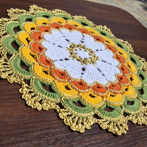 Puede incluir: Un tapete de ganchillo redondo y multicolor que presenta un patrón floral central blanco rodeado por anillos concéntricos de hilo naranja, amarillo y verde salvia. El borde exterior está acabado con un ribete decorativo festoneado en oro.
