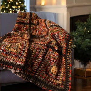 May include: A crocheted blanket in autumnal shades of brown, orange, and green. The blanket, with its square pattern and scalloped edge, is draped over a blue sofa. This handmade throw offers a cosy feel, adding a touch of warmth and handcrafted charm to a living room.