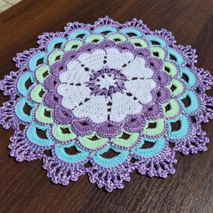 May include: A round, multi-colored crochet doily featuring a central white floral pattern surrounded by concentric rings of purple, light green, and bright turquoise yarn. The outer edge has a decorative, scalloped lace border in purple.