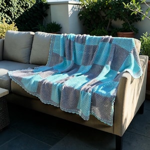 Può includere: Una coperta all'uncinetto fatta a mano con un motivo a scacchiera di quadrati alternati in azzurro e grigio. Il tessuto presenta un bordo smerlato ed è drappeggiato su un divano di colore neutro, mettendo in mostra la lavorazione a maglia aperta.