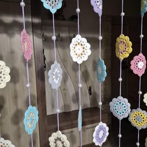 May include: A decorative hanging room divider featuring multiple vertical strands of light purple yarn. Each strand is adorned with round, multi-colored crocheted floral motifs and small white beads spaced evenly between the flower patterns.