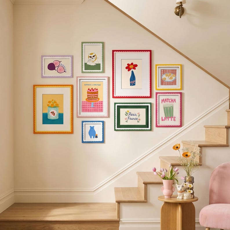 May include: A gallery wall arrangement featuring nine colorful framed prints with scalloped edges. The artwork includes illustrations of a cake, a flower in a vase, a matcha latte, a blue bear, a cat, fruit, and text reading Paris, France.
