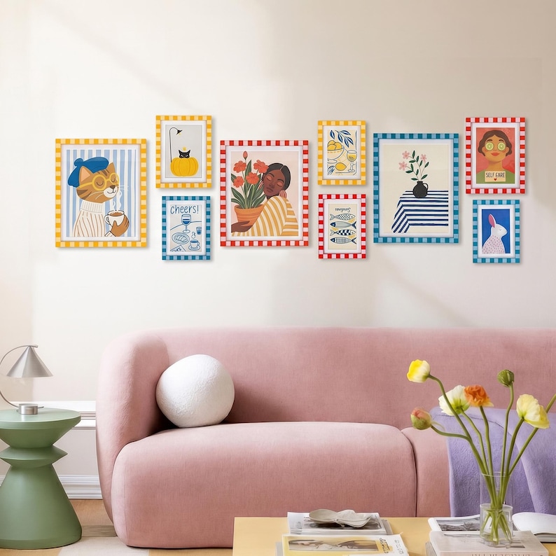 May include: A collection of framed art prints with colorful illustrations, including a cat, a person holding flowers, and fish. The frames have yellow, red, and blue borders. The art is displayed on a white wall above a pink sofa.