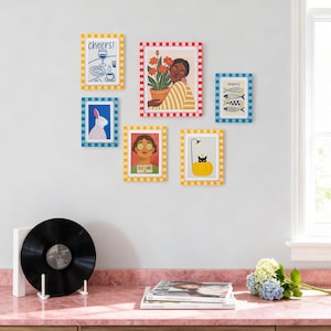 May include: A collection of framed art prints with colorful illustrations. The prints feature images of a person with flowers, a rabbit, fish, a cat, and text that says "Cheers!" and "Self Care". The frames have checkered borders.
