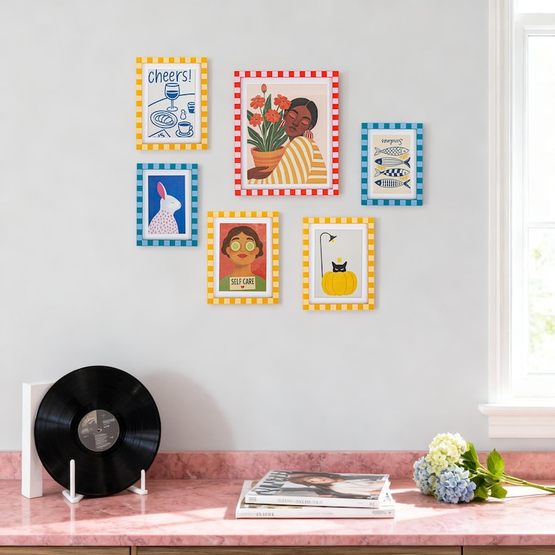 May include: A collection of framed art prints with colorful illustrations. The prints feature images of a person with flowers, a rabbit, fish, a cat, and text that says "Cheers!" and "Self Care". The frames have checkered borders.