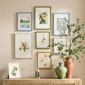 May include: A gallery wall featuring framed botanical prints and landscape art in various sizes and colors. Frames include blue, green, white, and yellow with beaded edges. A wooden cabinet holds a vase, a green vase, and candles.