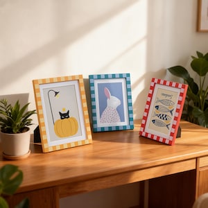 May include: Three framed art prints on a wooden surface. The frames are yellow, blue, and red with checkered patterns. The prints feature a black cat in a pumpkin, a white rabbit, and fish with the word "Sardines".