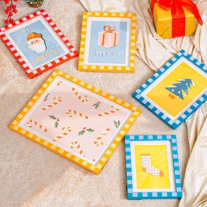 May include: A collection of framed Christmas-themed art prints. The frames are in various colors, including red, yellow, and blue, with a checkered pattern. The prints feature festive images like Santa Claus, candy canes, a gift box, a Christmas tree, and a sock. The word "CELEBRATE" is visible.