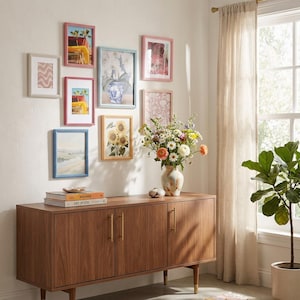 Puede incluir: Una colección de obras de arte enmarcadas en varios tamaños y colores, dispuestas en una pared blanca. Los marcos son en tonos rosa, azul y blanco, con detalles de cuentas. Un mueble de madera con tiradores dorados y un jarrón de flores completan la decoración.