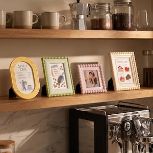 May include: Four decorative picture frames in yellow, green, pink, and cream colors displayed on a wooden shelf. The yellow frame is oval with a Spirit Cafe menu, the green frame is rectangular with coffee beans, the pink frame holds a photo, and the cream frame displays a dessert card.