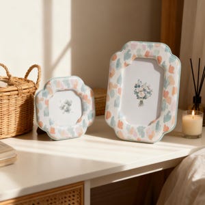 May include: Two decorative picture frames with a floral design. The frames have a scalloped edge and are decorated with pastel-colored brushstrokes. The frames are on a white surface, with a wicker basket and a lit candle in the background.