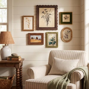 May include: A gallery wall featuring six framed botanical and landscape prints in various shapes and sizes, arranged above a striped armchair with a green throw blanket, next to a wooden side table with a lamp and stacked books.