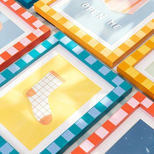 May include: A collection of colorful framed art prints. The frames feature a variety of patterns, including checkered and square designs in shades of orange, teal, and yellow. One print displays a sock illustration on a yellow background.